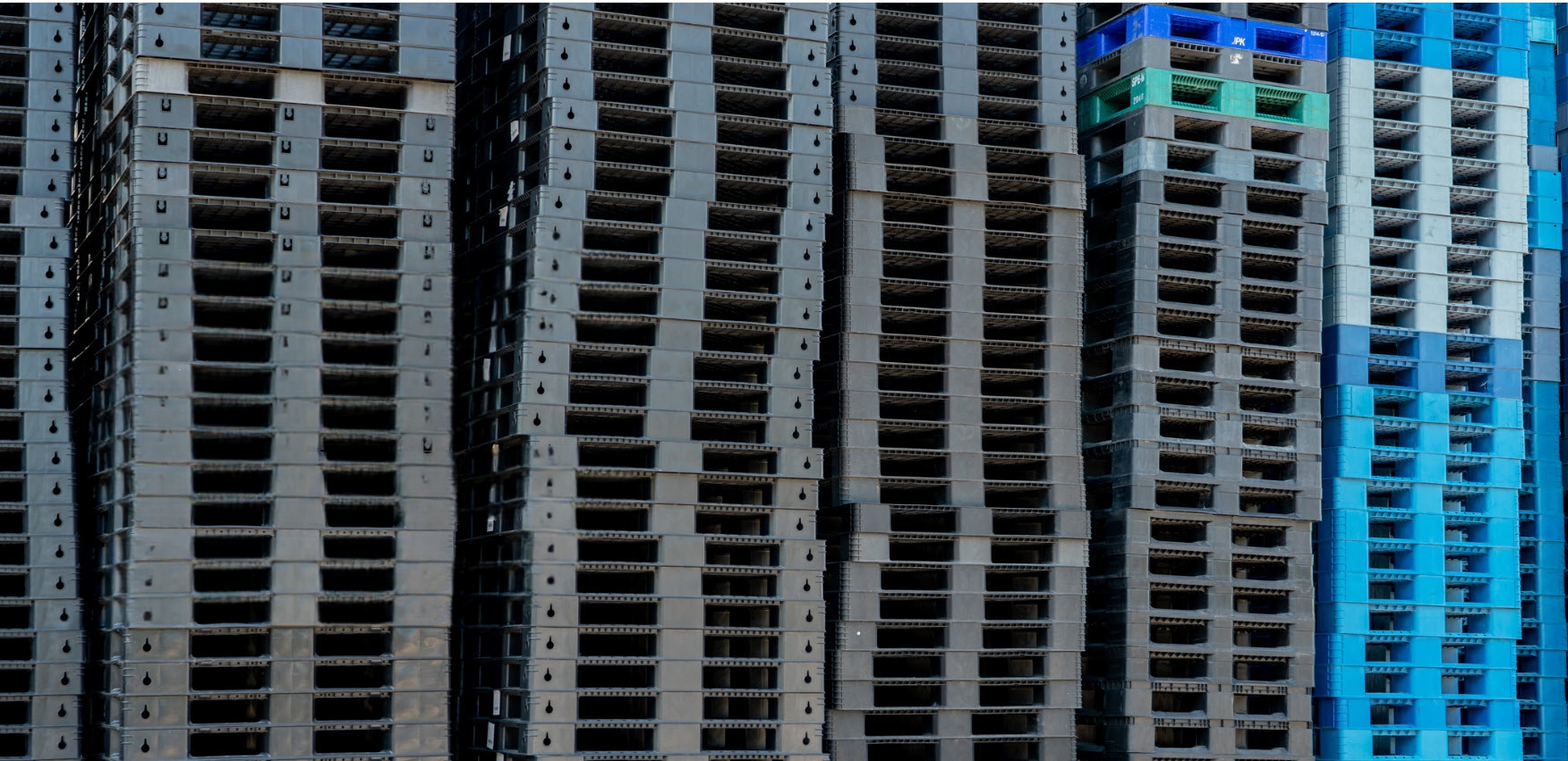 Composite logistics pallets in gray, blue, green, and white stacked in tall columns at a staging yard, ready for shipment